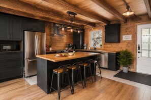 A modern kitchen with a large island, dark cabinetry, stainless steel appliances, and a wooden ceiling. - Spa, Pool Table & Nature | Les Bons Vivants (Saint-Côme)