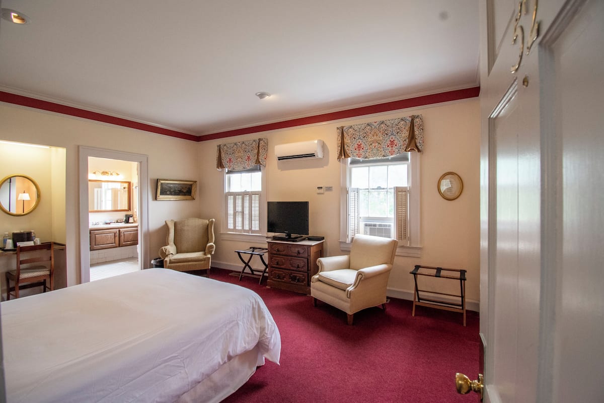 Una habitación de hotel con una cama grande, dos sillones, un televisor, un aparador y un equipo de aire acondicionado.