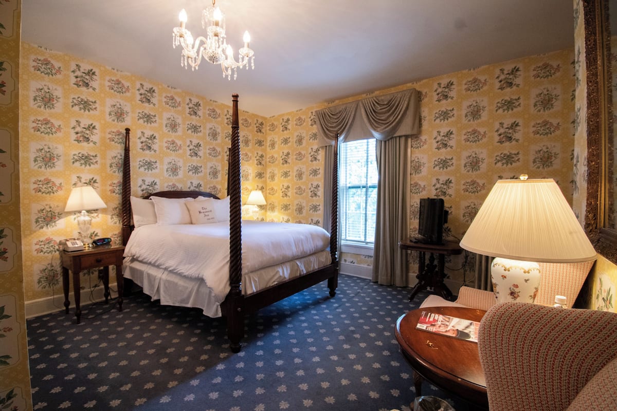 Una cama con dosel, dos lámparas de noche, una mesita de madera, una silla, una mesa, una ventana con cortinas y un candelabro.