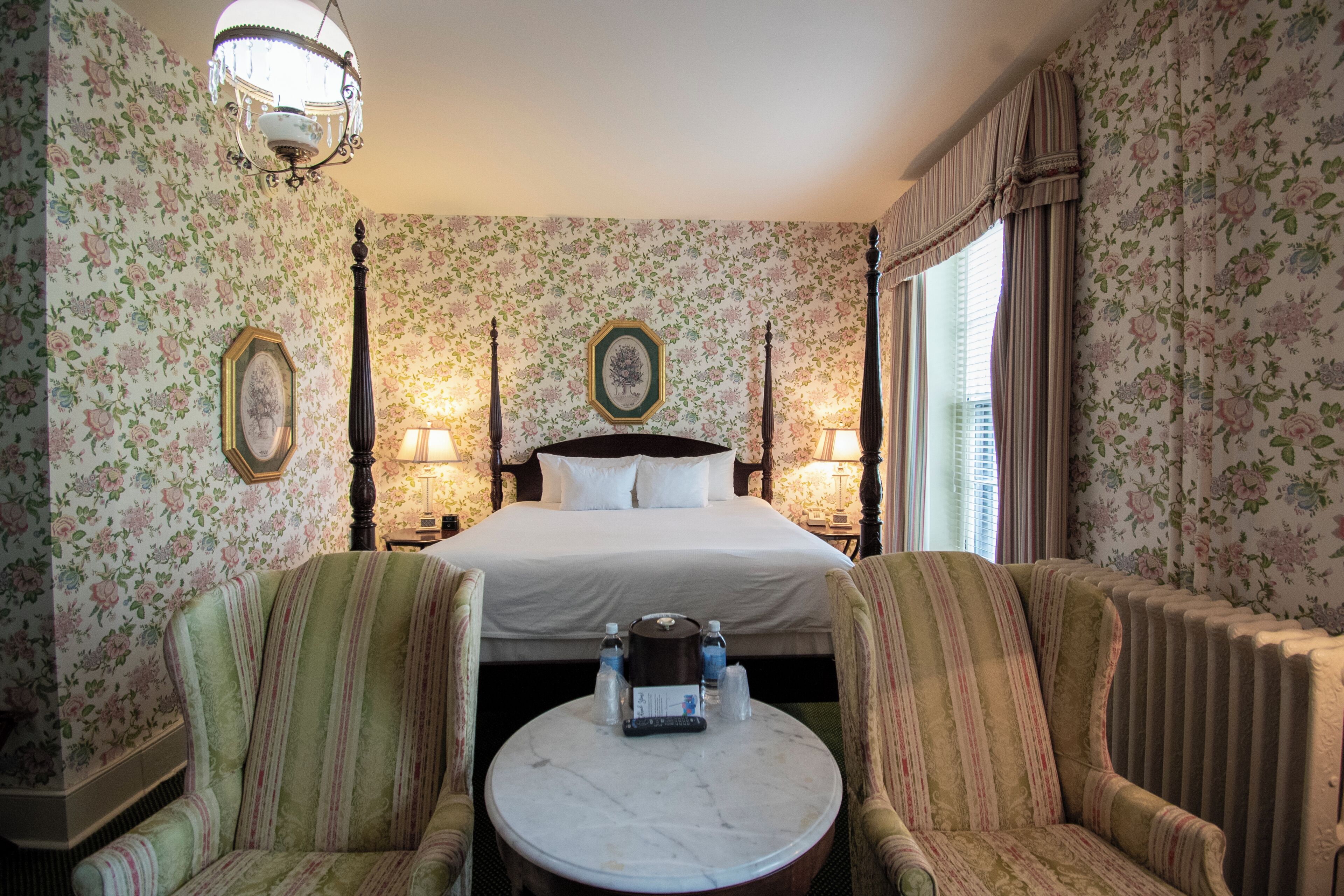A hotel room with a bed, two armchairs, a round table, a chandelier, and floral wallpaper.