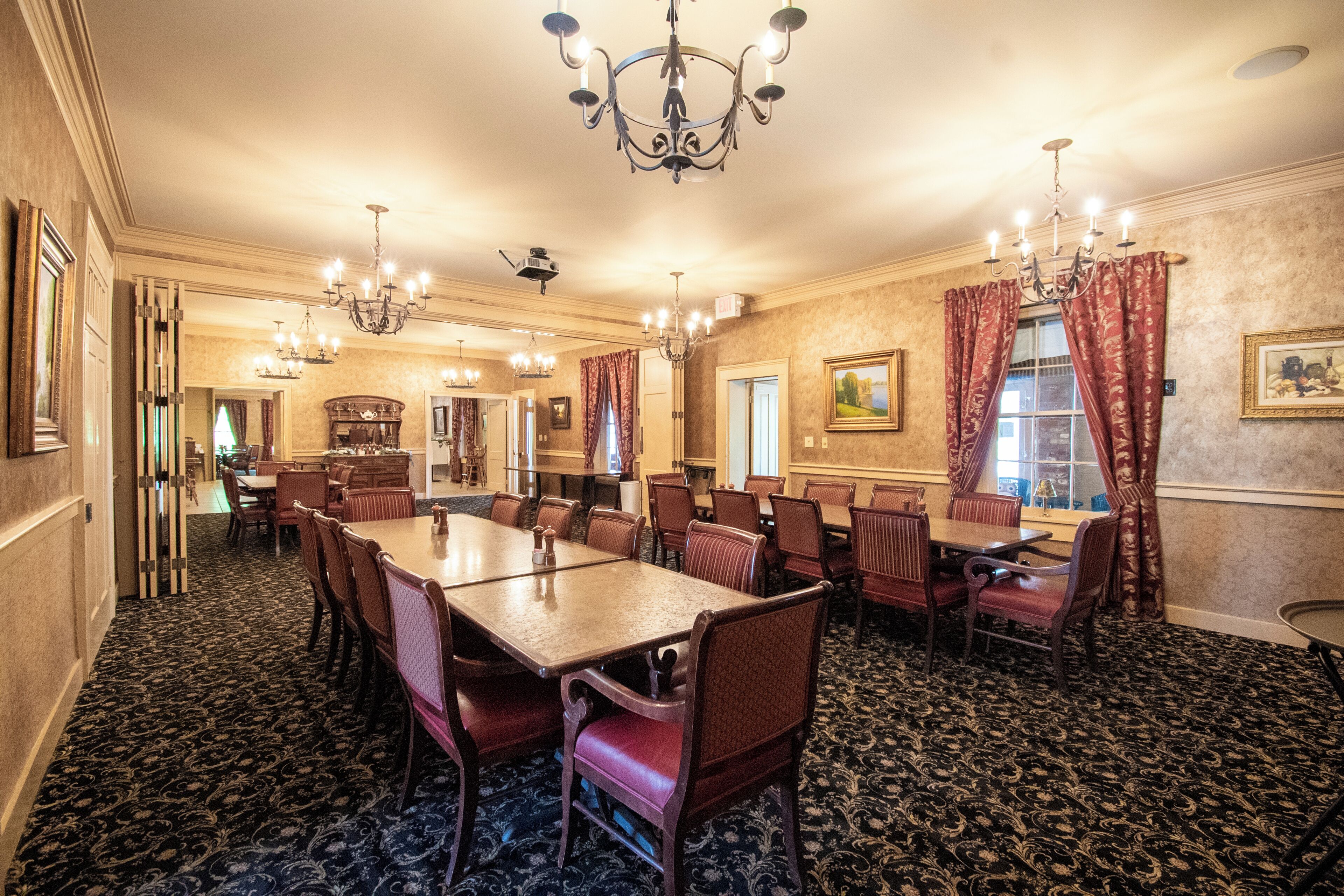 A formal dining room with a large table, multiple chairs, chandeliers, and patterned carpet.