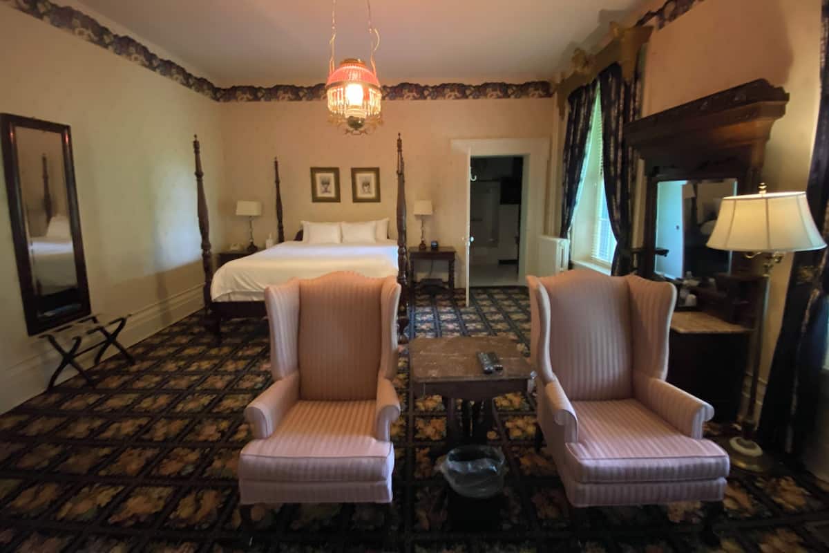 Una habitación de hotel con una cama con dosel, dos sillones, una mesita, una araña y un espejo.