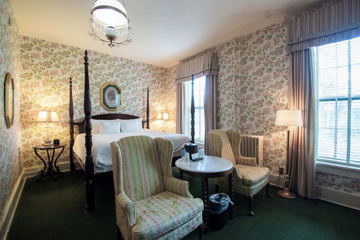 Una habitación de hotel con papel tapiz floral, una cama con dosel, dos sillones, una mesa redonda y un candelabro.