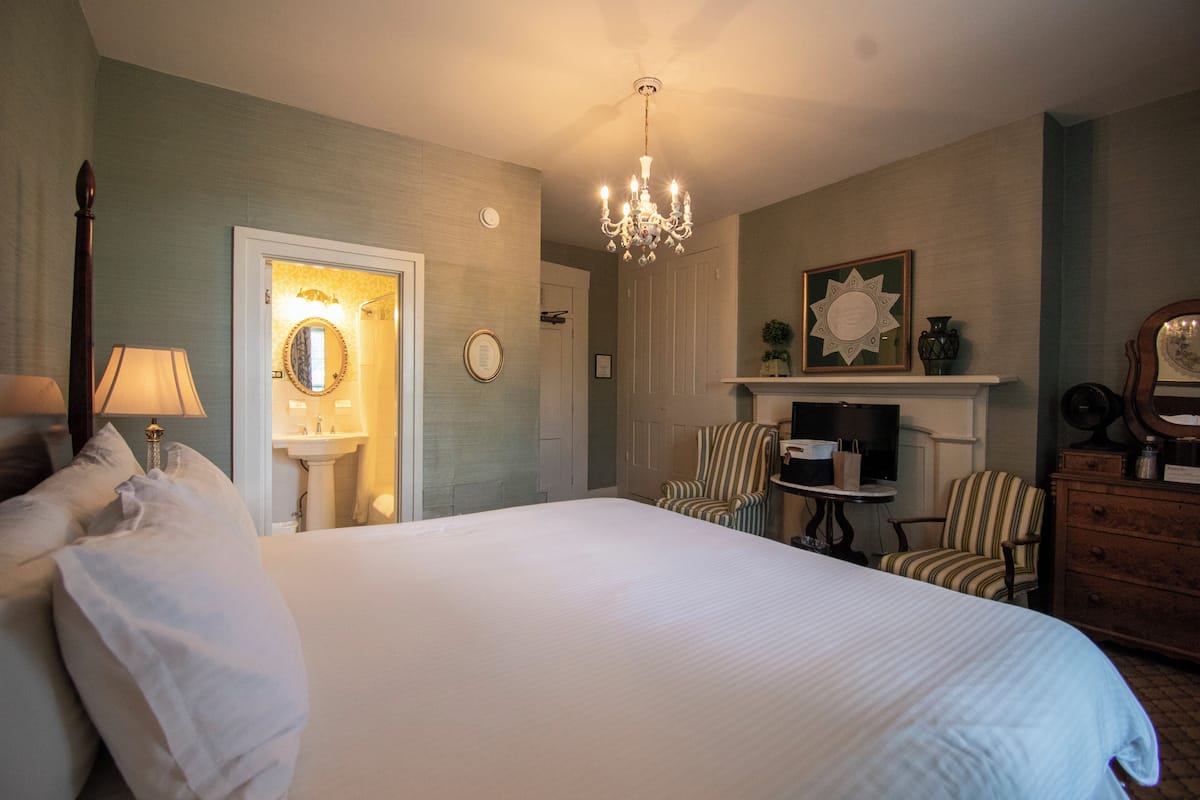 Una habitación de hotel con una cama grande, una araña, una chimenea, un espejo y un sillón a rayas.