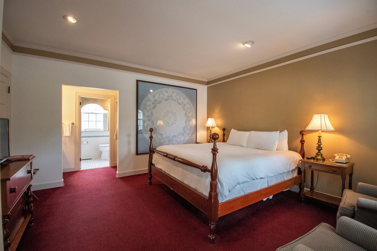Habitación de hotel con una cama grande, lámparas de noche, una mesita de noche de madera, una silla, un cuadro en la pared y un baño visible a través de una puerta abierta.