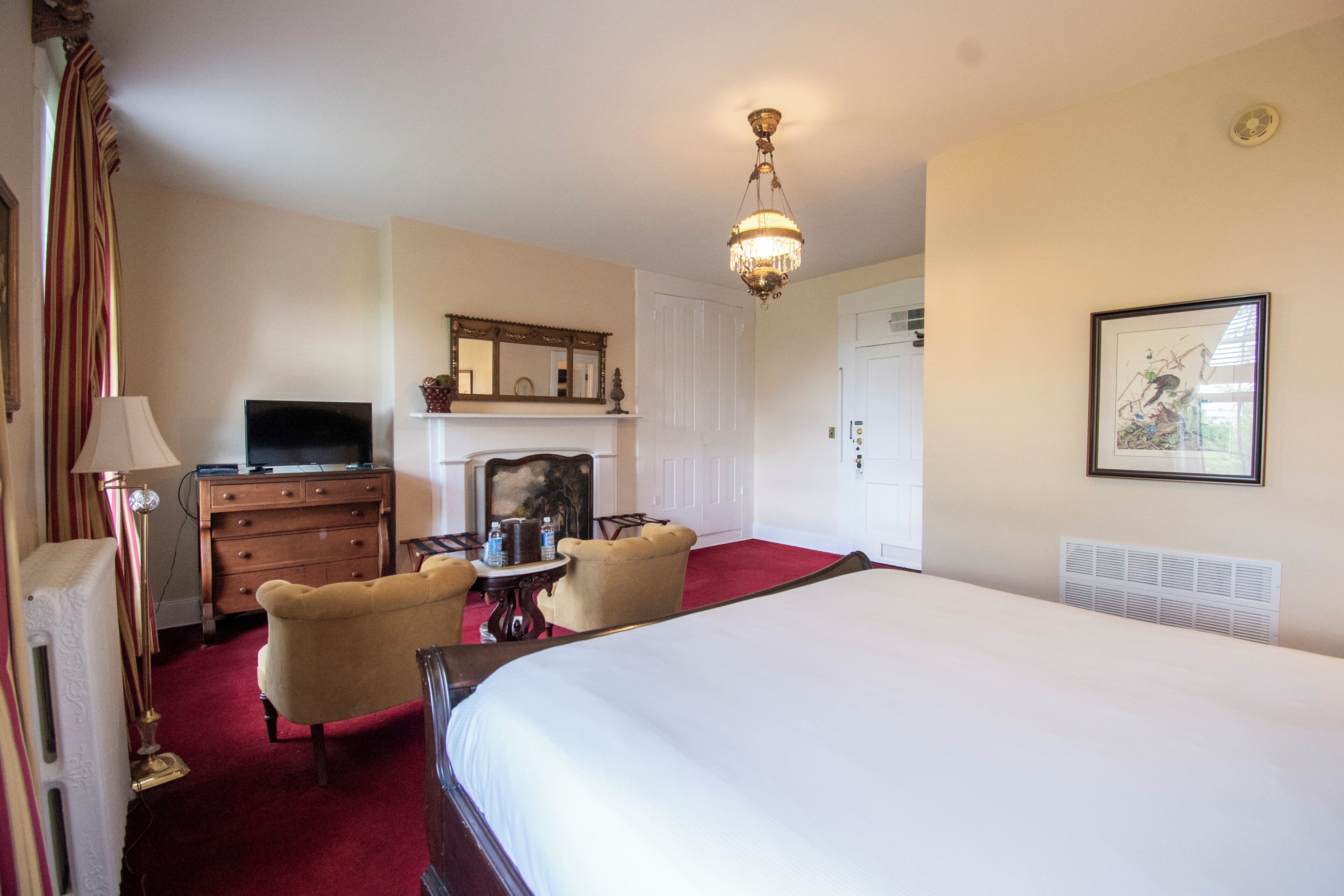 A hotel room with a large bed, a television, a dresser, a chair, a table, and a chandelier.