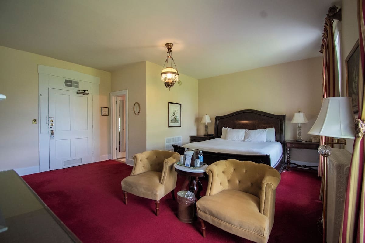 Habitación de hotel con cama, dos sillones, una mesita, un cubo de basura, una araña y una puerta.