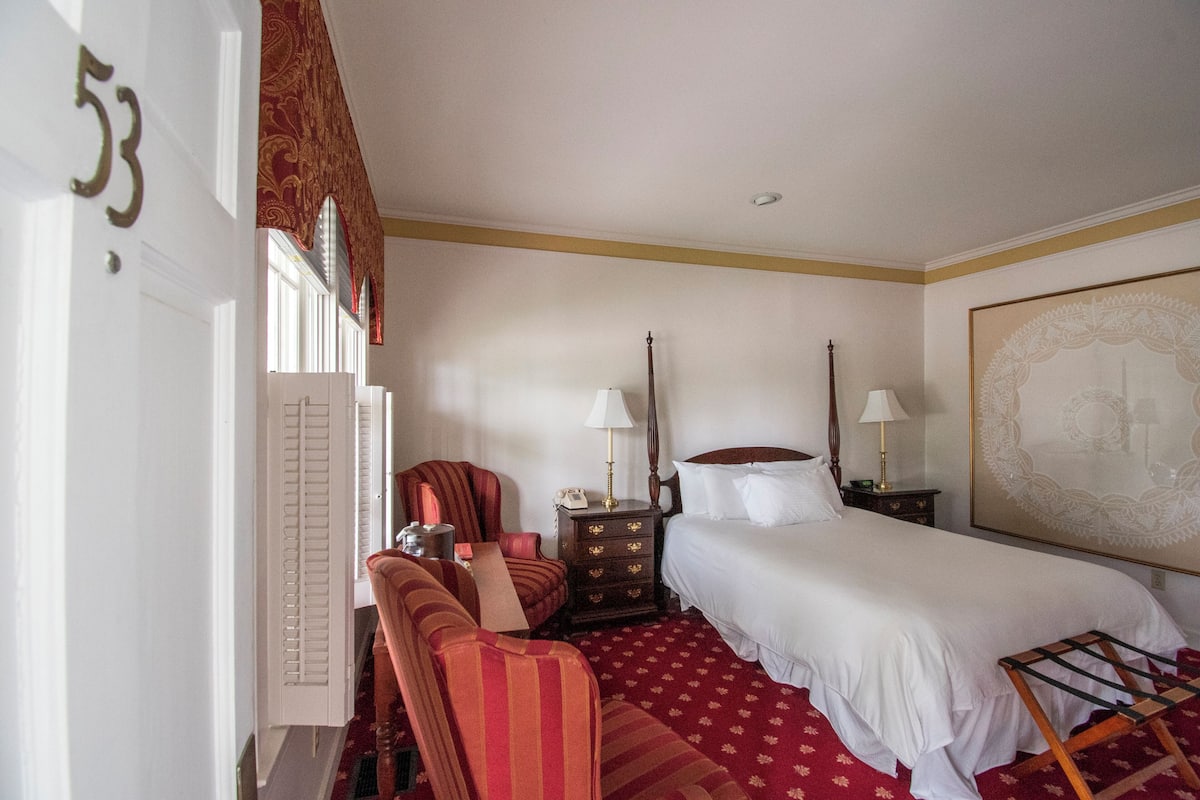 Habitación de hotel con cama, una silla, una mesita de noche, una lámpara y una obra de arte grande en la pared.