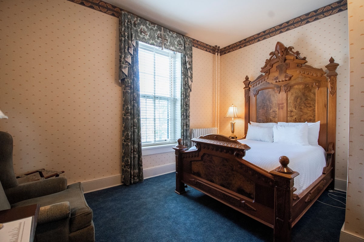 Un dormitorio tradicional con cama con dosel, una silla y una ventana con cortinas.