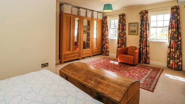 A bedroom with a bed, a wooden bench, a wardrobe, a chair, a red rug, and a window with curtains. - Ormiston House (Pewsey)