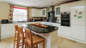 A kitchen with white cabinets, a central island with chairs, a refrigerator, and a window with curtains. - Ormiston House (Pewsey)