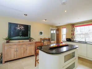 A kitchen with a central island, wooden cabinets, a dining area, and a window with curtains. - Ormiston House (Pewsey)