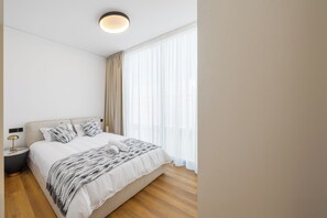 Ein modernes Schlafzimmer mit einem großen Bett, einem Nachttisch, einer Lampe und einem Fenster mit transparenten Vorhängen.