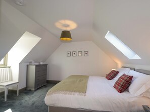 Ein Schlafzimmer mit schrägen Wänden, einem Bett mit weißer Bettwäsche, einer Bank, einem Schminktisch und einem Dachfenster.
