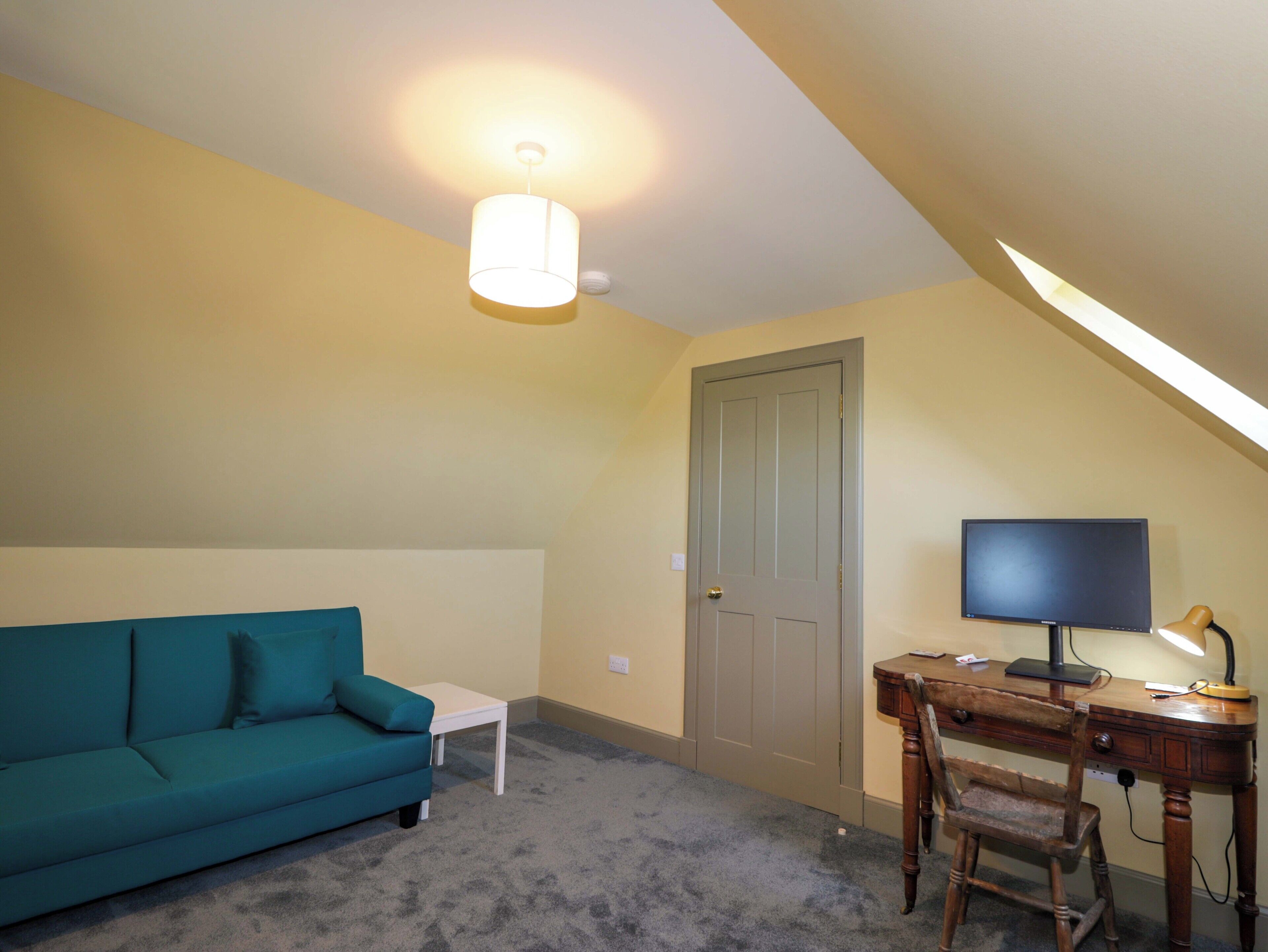 A room with a teal sofa, a small white table, a wooden desk with a computer, and a door.