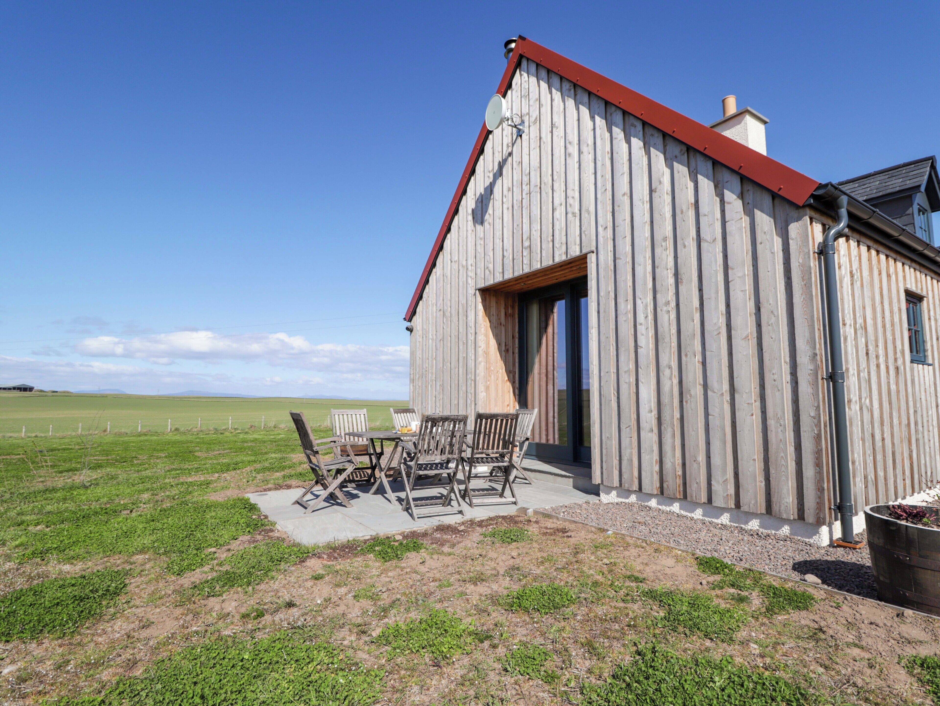 Une grange en bois avec un toit rouge, une terrasse avec des chaises et une vue sur un champ verdoyant.