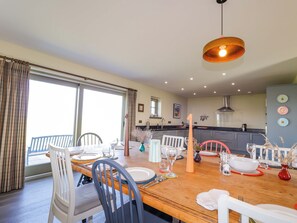A dining area with a wooden table set for four, chairs, a hanging light fixture, and a view of the kitchen in the background. - The Mandrel (Tain)