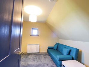 Ein Zimmer mit einem blauen Sofa, einem weißen Tisch, einem Heizkörper, einem kleinen Fenster und einer Tür.