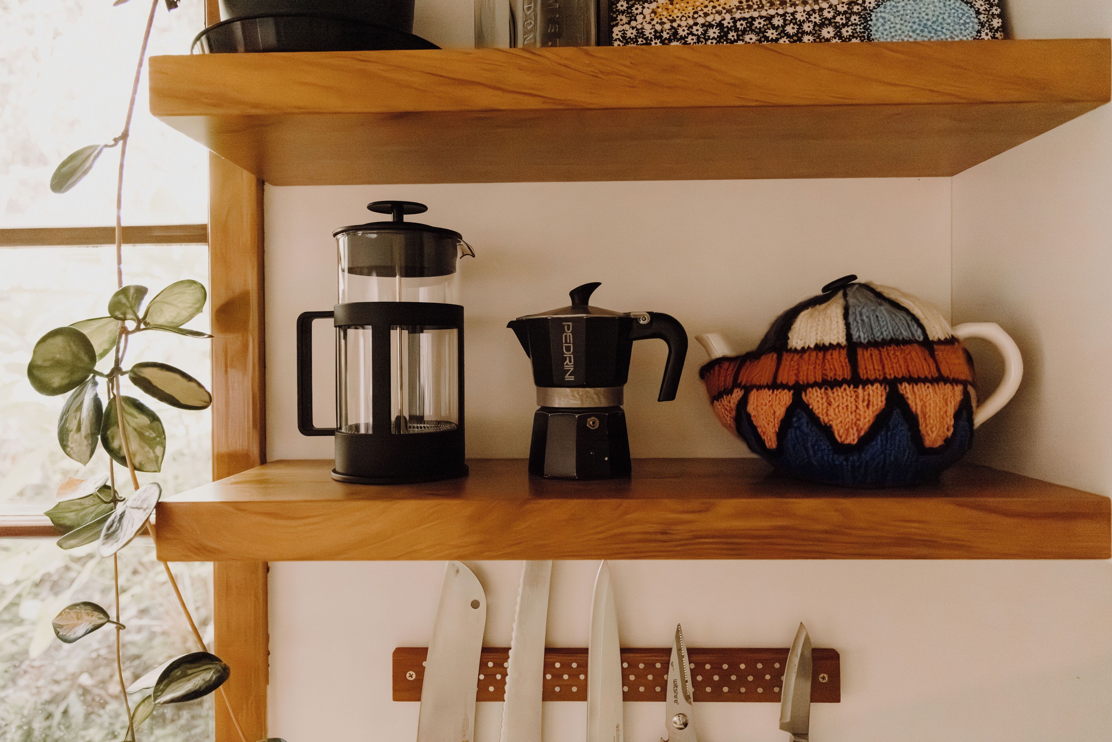 Una estantería con una cafetera, una tetera y una taza tejida.
