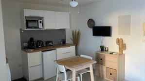 A compact kitchen with a microwave, coffee maker, toaster, and a small refrigerator. There's a wooden table with two chairs, a ceiling fan, and a flat-screen TV mounted on the wall. - STUDIO LE FLAMANT 20M FROM THE BEACH (Palavas-les-Flots)
