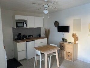 Eine kompakte Küche mit Mikrowelle, Kaffeemaschine, Toaster und kleinem Kühlschrank. Ein Holztisch mit zwei Stühlen, ein Deckenventilator und ein Flachbildfernseher an der Wand sind ebenfalls vorhanden.