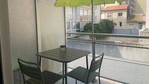 A balcony with a table and chairs, a green umbrella, and a view of buildings and greenery. - STUDIO LE FLAMANT 20M FROM THE BEACH (Palavas-les-Flots)
