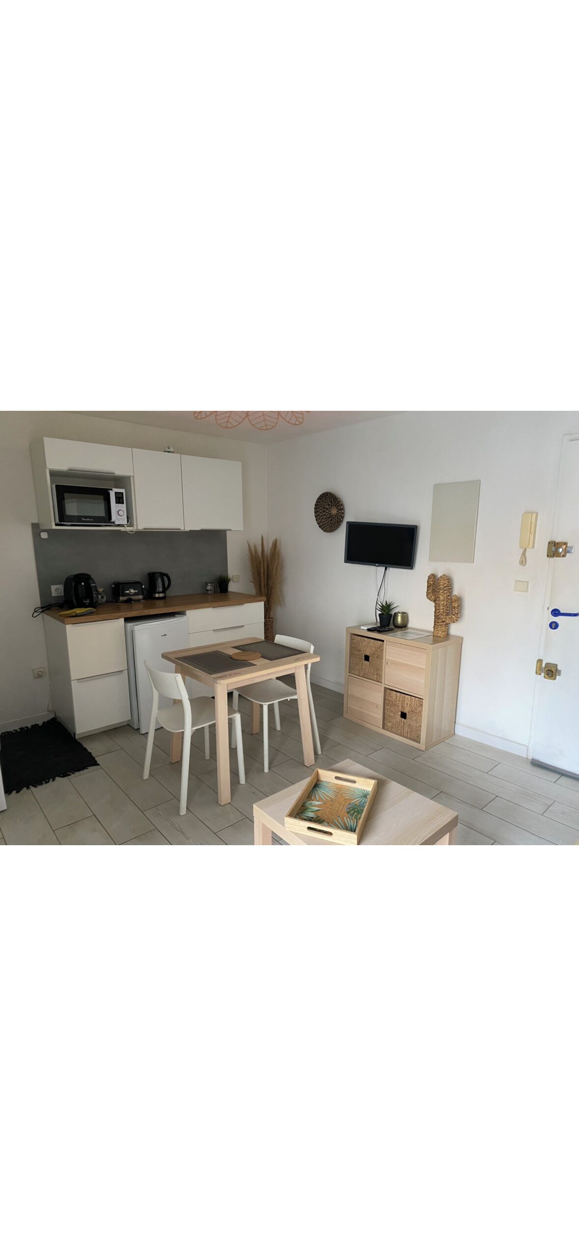 A compact kitchen with a microwave, toaster, and coffee maker. There is a dining table with chairs and a small cabinet with drawers. A flat-screen TV is mounted on the wall, and a door with a handle is visible.