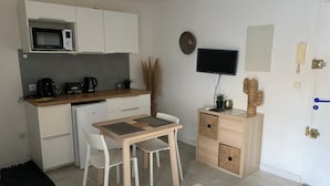A compact kitchen with a microwave, toaster, and coffee maker. There is a dining table with chairs and a small cabinet with drawers. A flat-screen TV is mounted on the wall, and a door with a handle is visible. - STUDIO LE FLAMANT 20M FROM THE BEACH (Palavas-les-Flots)