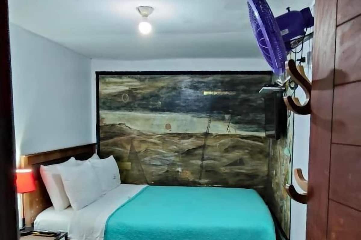 Une chambre à coucher avec un lit, une table de chevet, un ventilateur fixé au mur et une porte.