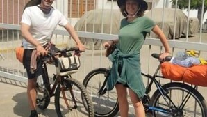 Een man en een vrouw staan naast fietsen voor een metalen hek met een gebouw op de achtergrond.