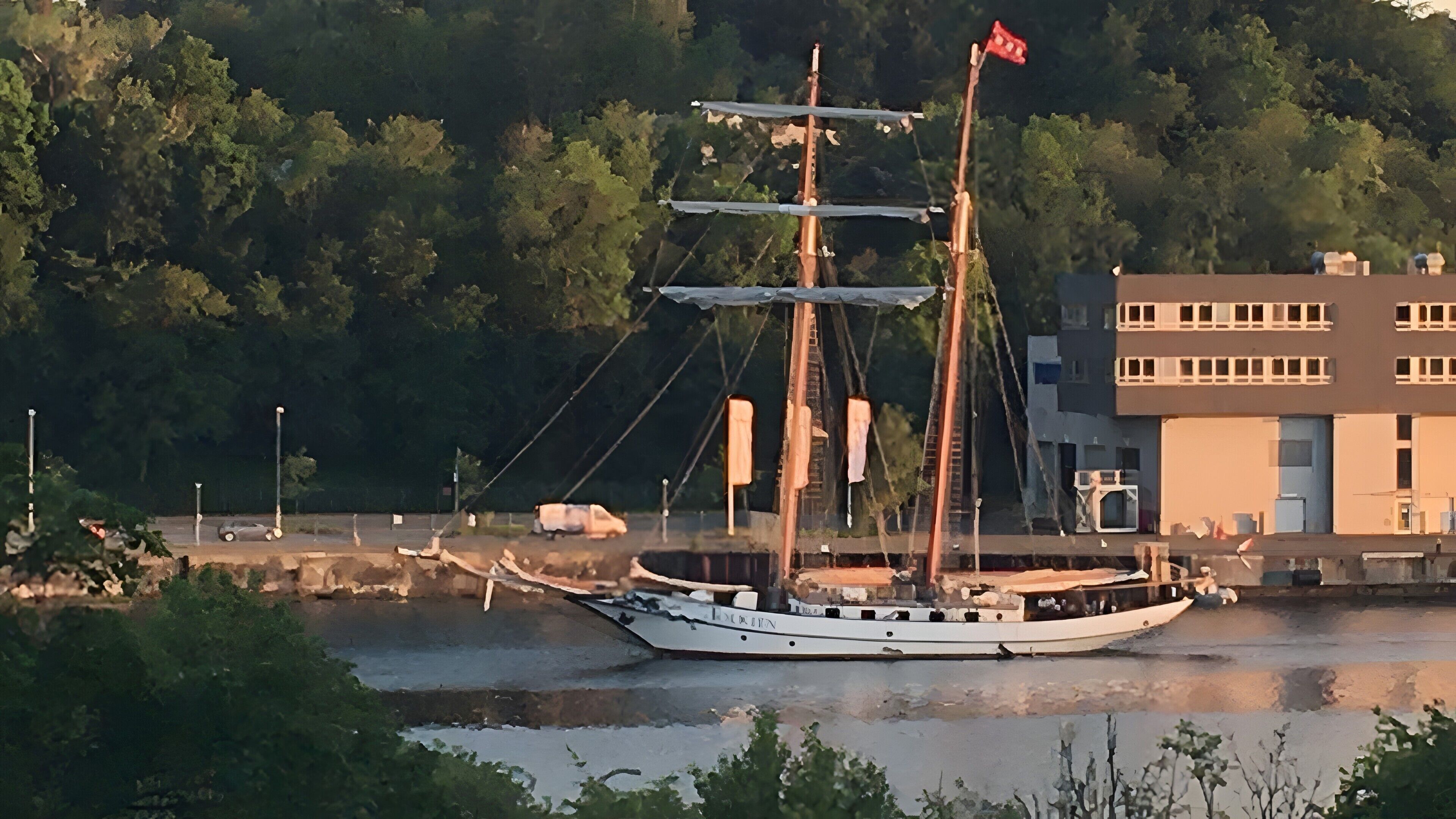 A boat with a flag, docked near a building with multiple windows.