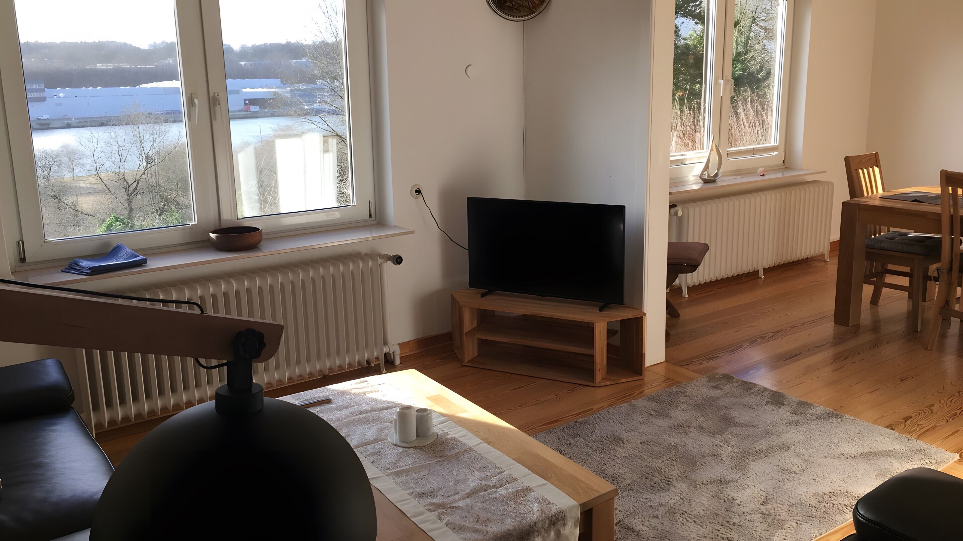 A living room with a TV, wooden furniture, a radiator, and large windows with a view of a body of water.