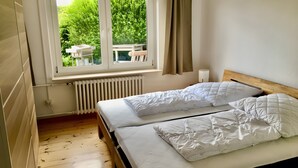 Et soveværelse med to senge, et vindue der ud til grønne omgivelser, en radiator og et natbord med en lampe.