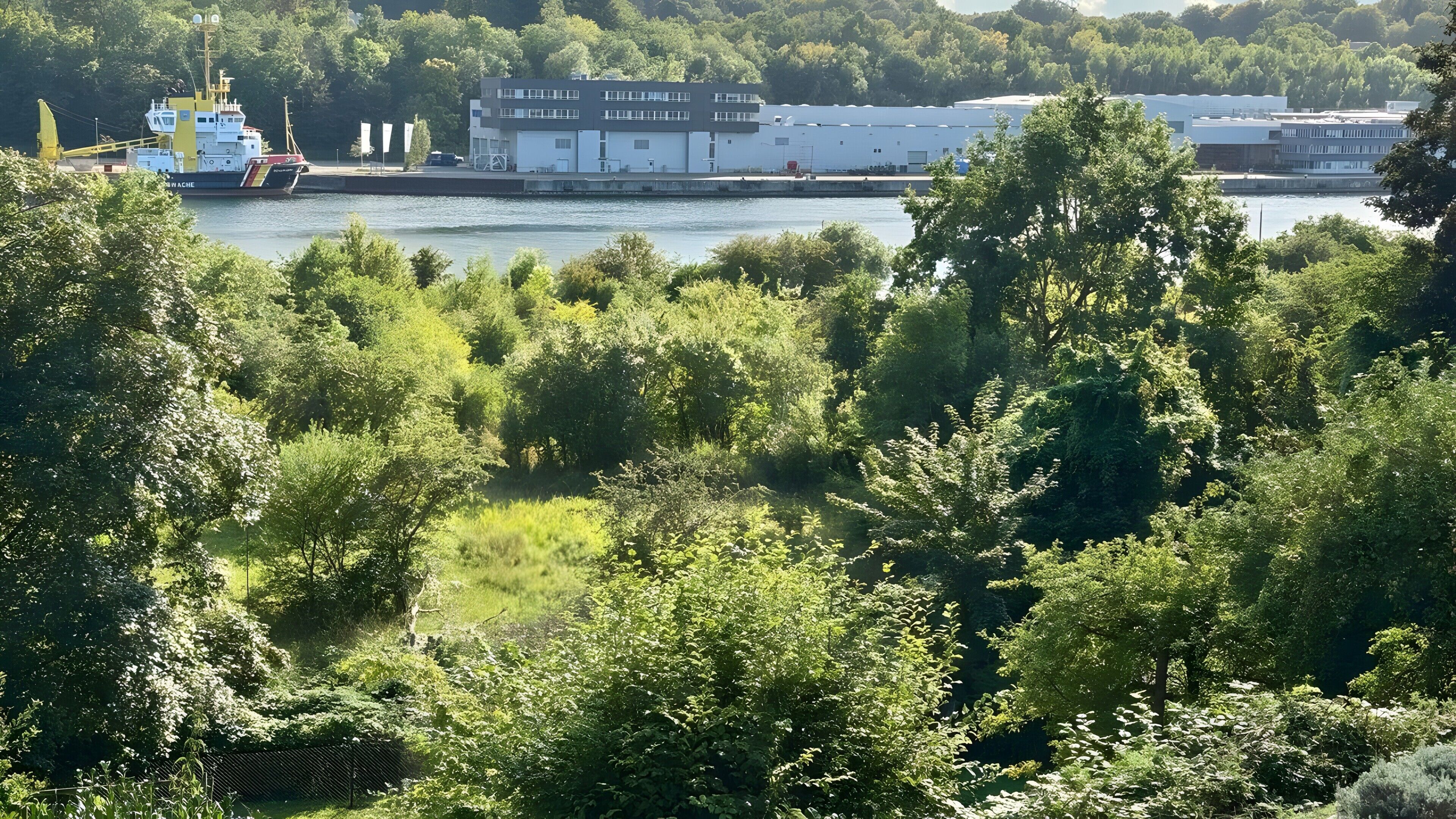 A river with a ship docked, a modern building with a flat roof, and dense greenery in the foreground.