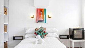 A hotel room with a bed, white walls, a painting, a television, and a small table. - Oriental Eight | By My Getaways (Brighton)