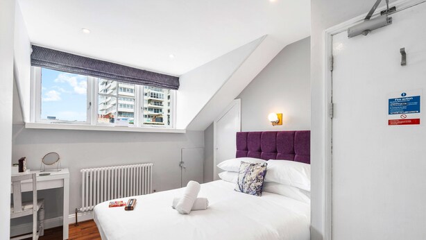 A bedroom with a bed, a window, a radiator, a bedside table, and a door. - Oriental Nine | By My Getaways (Brighton)