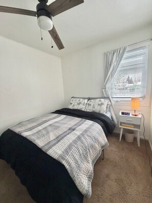 A bedroom with a bed, a nightstand with a lamp, a ceiling fan, and a window with curtains.