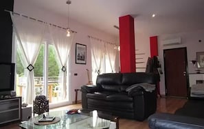 Un salon avec un canapé en cuir noir, une table basse en verre et un téléviseur. Des rideaux sont accrochés aux fenêtres, et on peut apercevoir un pilier rouge.