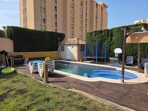 Une piscine avec un petit espace aménagé, des chaises longues et un panier de basket.