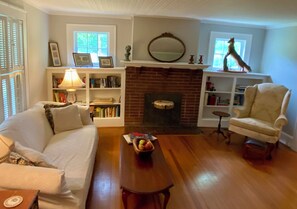 Uma sala de estar com lareira, uma poltrona branca, uma mesa de centro de madeira e uma estante de livros.