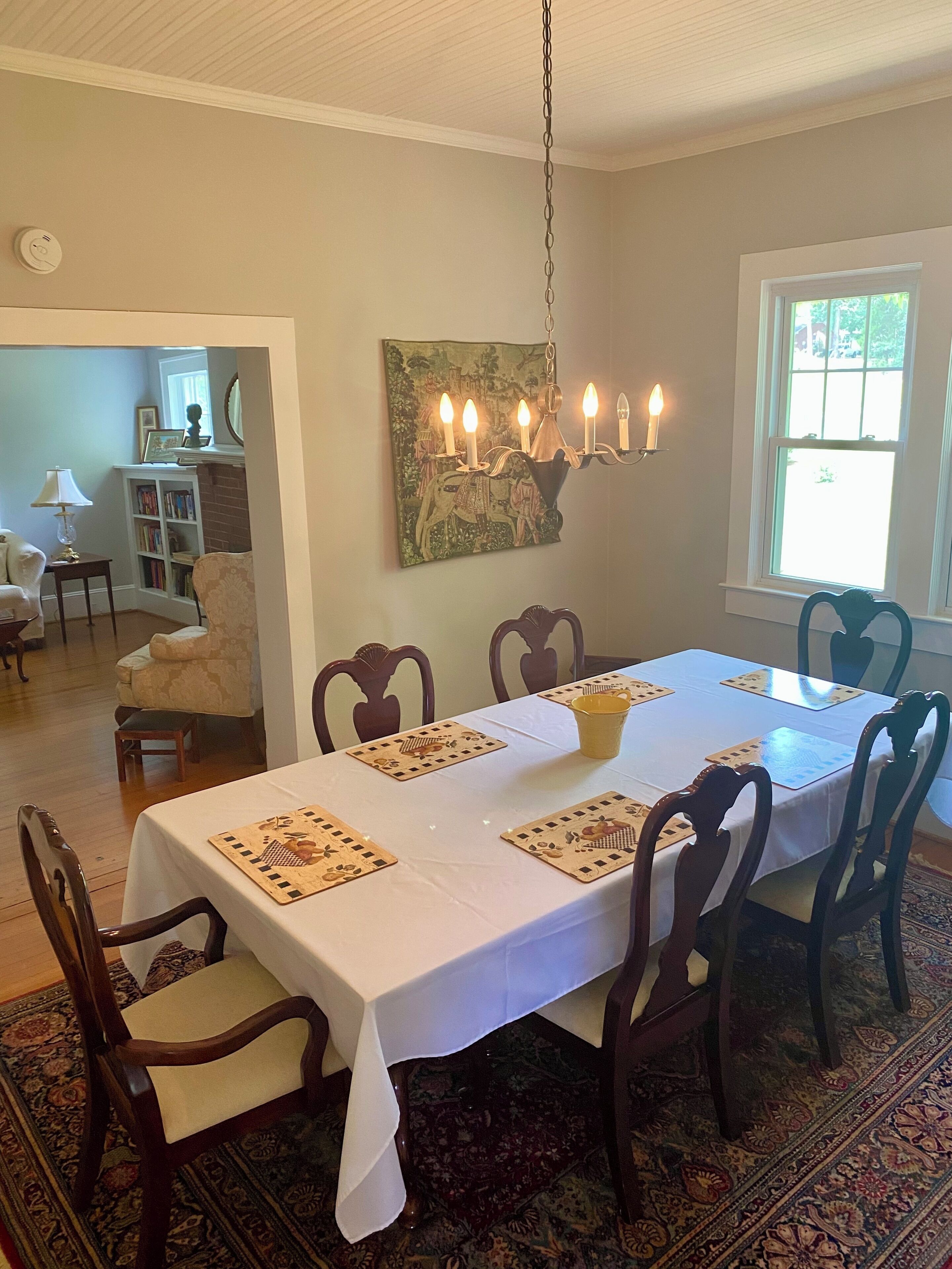 A dining room with a long table, chairs, a chandelier, and a bookshelf.