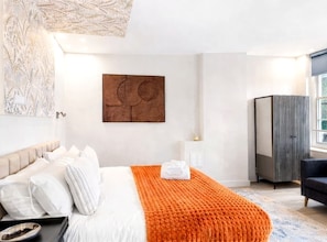 A hotel room with a bed, a large orange throw blanket, a wall-mounted artwork, a wardrobe, and a window. - Luxury Apartment Ideal For Contractors & Couples (Portsmouth)