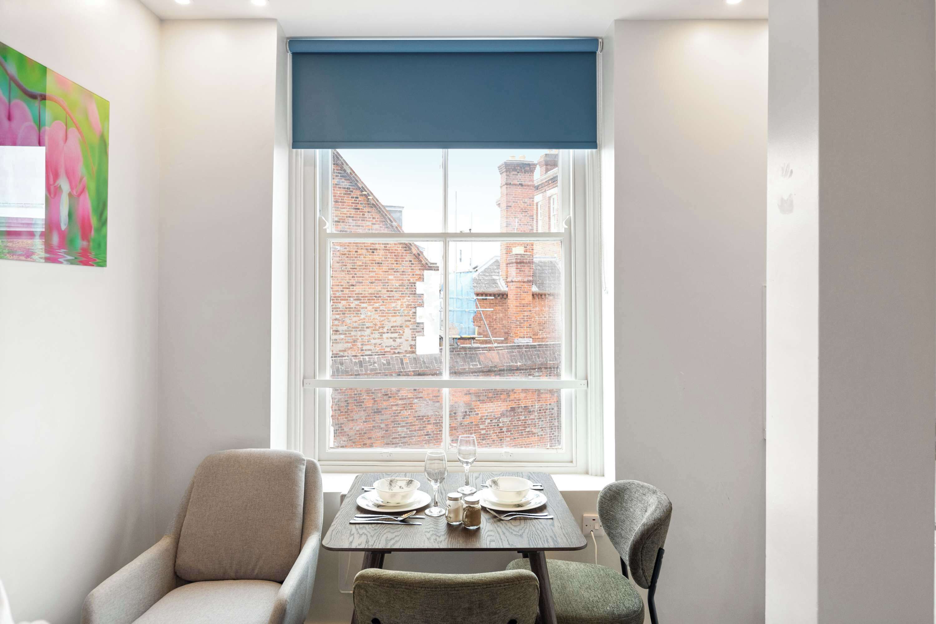 A corner room with a dining table, two chairs, and a view of brick buildings through a window with a blue blind.