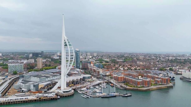 Een modern gebouw met een opvallende spits, een jachthaven met boten, en een stadsgezicht op de achtergrond.