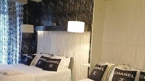 A hotel room with two beds, each with white bedding and multiple pillows featuring the Chanel logo.