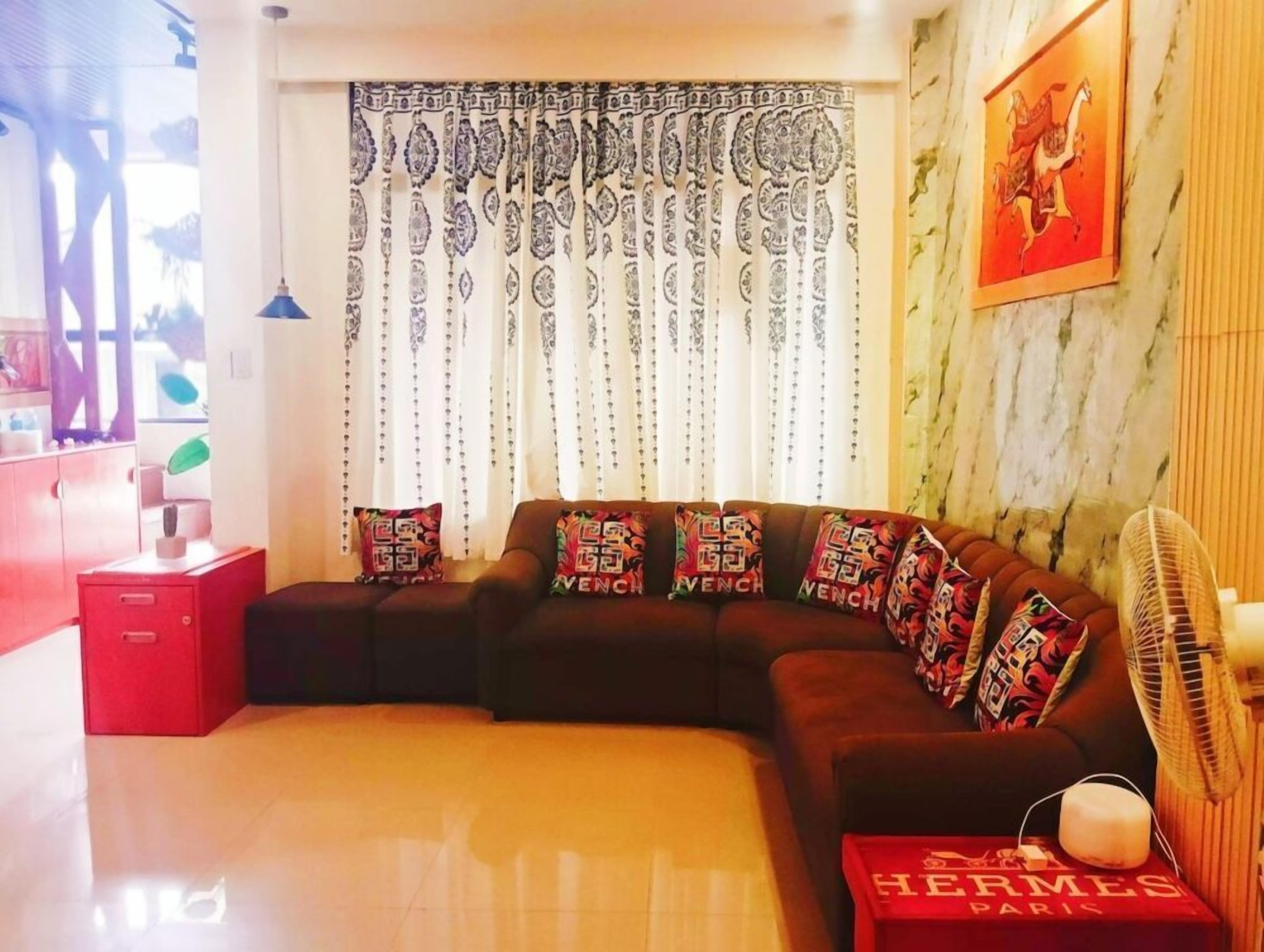Ein Wohnzimmer mit einem Ecksofa, gemusterten Vorhängen, einem an der Wand befestigten Kunstwerk und einer roten Kiste mit dem Aufdruck 'HERMES PARIS'.