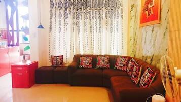 Ein Wohnzimmer mit einem Ecksofa, gemusterten Vorhängen, einem an der Wand befestigten Kunstwerk und einer roten Kiste mit dem Aufdruck 'HERMES PARIS'.