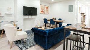 Ein modernes Wohnzimmer mit weißer Ziegelwand, einem flachbildfernseher über einem Kamin, einem Essbereich mit Holztisch und blauen Stühlen sowie einem blauen Samtsofa.