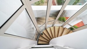 A spiral staircase with a wooden handrail and white walls, leading to a balcony with a view of greenery and a red building. - Avenue Palm (Eforie Nord)