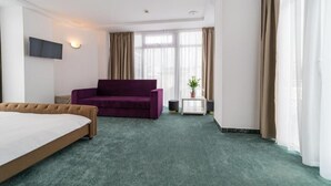 A hotel room with a bed, a purple sofa, a small table with a plant, and a TV mounted on the wall. - Avenue Palm (Eforie Nord)